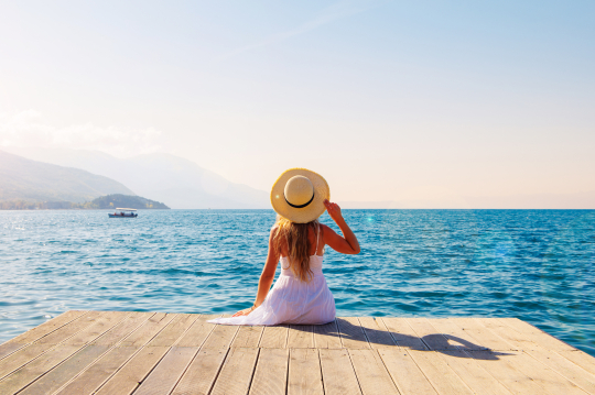 Frau mit Hut sitzt auf einem Steg und Blickt auf das Meer, im Hintergrund Berge und das Meer